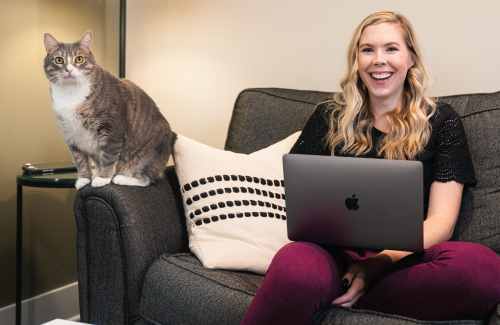 Megan works on her couch while her cat sits on the arm rest next to her